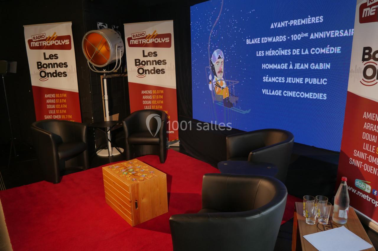 Studio avec fauteuils noirs, table basse en bois, écran affichant un programme d'événements et panneaux publicitaires.
