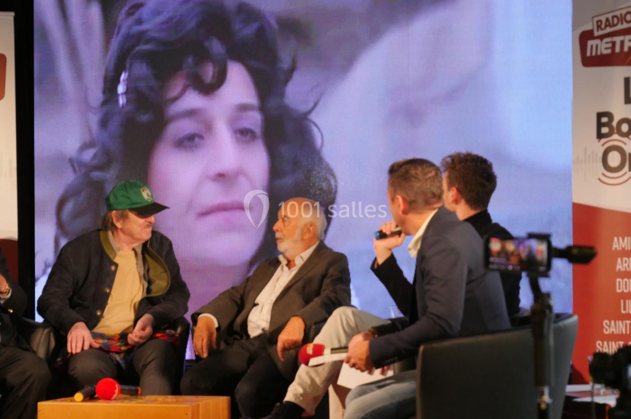 Des personnes discutent sur une scène avec un grand écran en arrière-plan affichant un visage féminin.