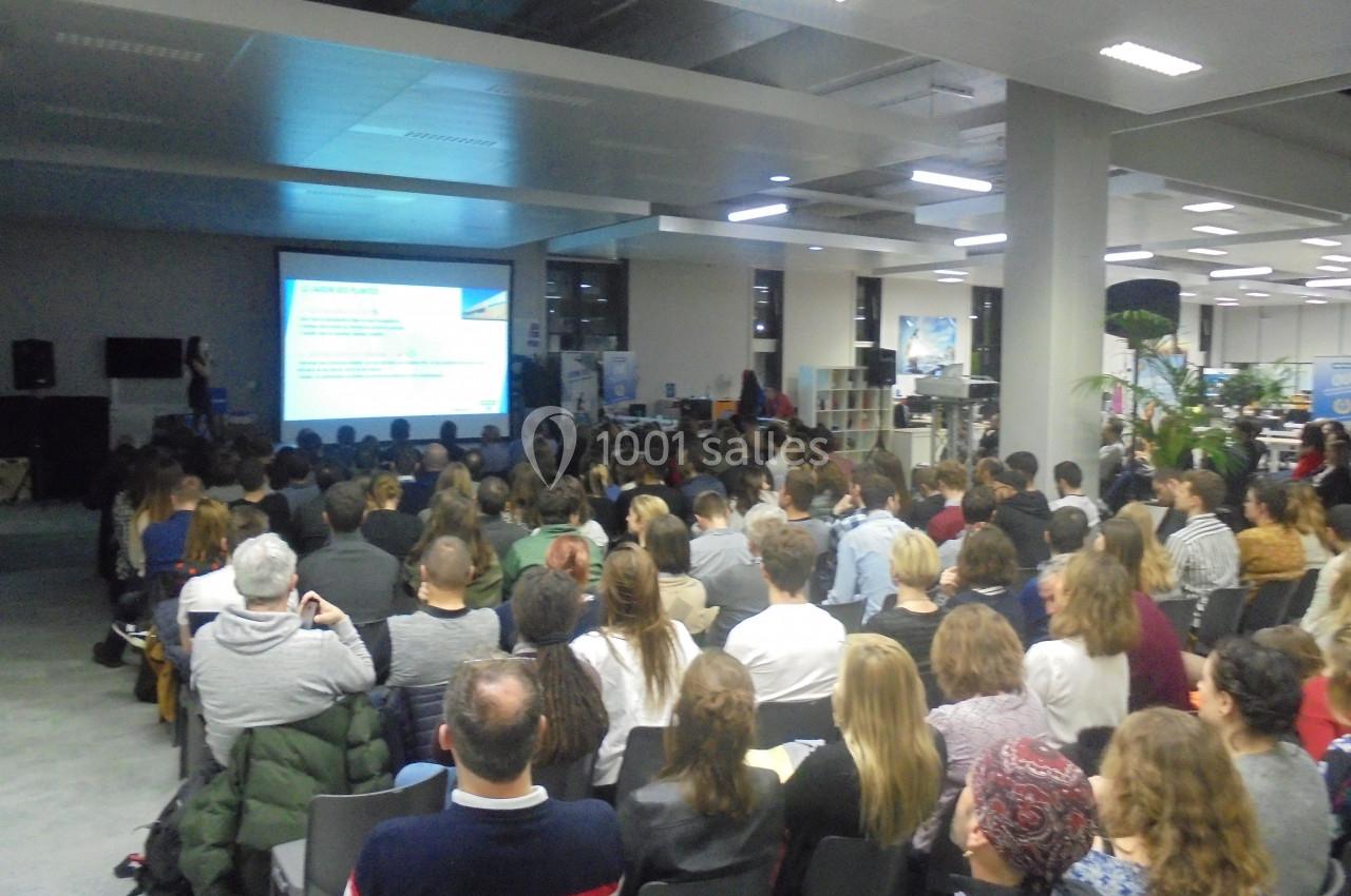 Salle comble avec des participants assis, assistant à une présentation projetée sur un écran dans un espace moderne.
