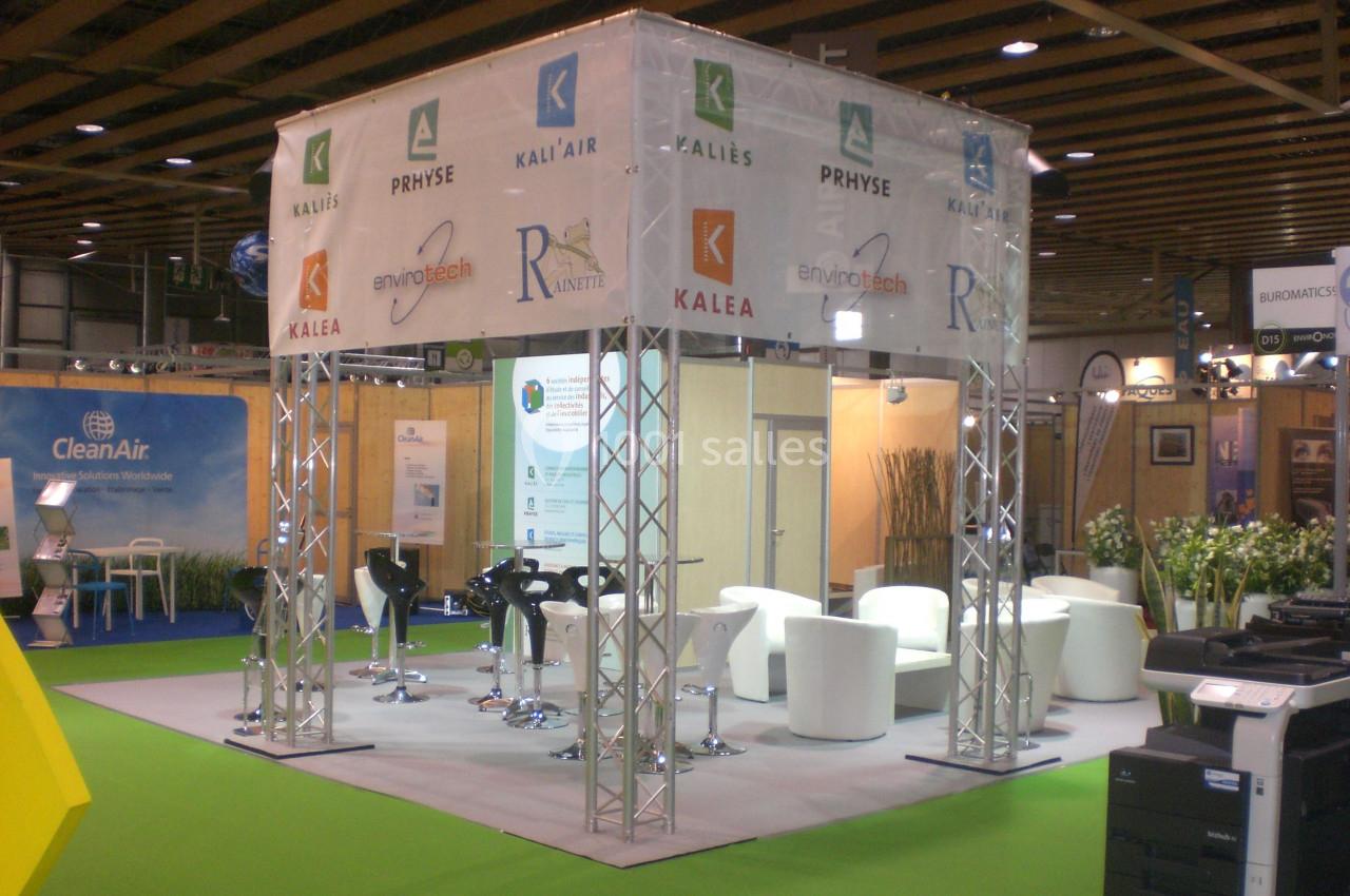 Stand d'exposition avec des structures métalliques, des logos d'entreprises, des chaises hautes et des fauteuils blancs.