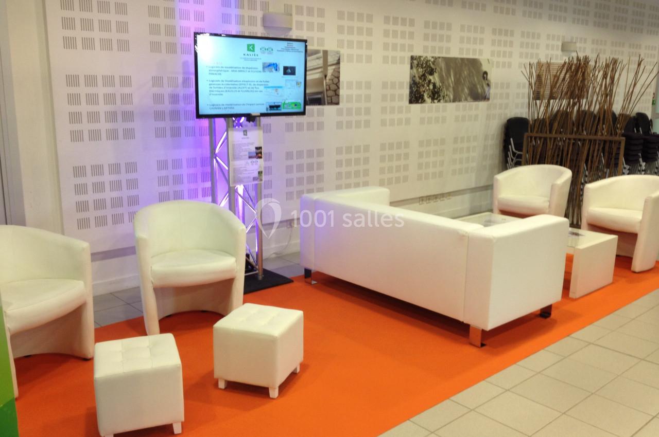 Espace d'attente avec fauteuils et canapé blancs, table basse et écran sur pied, posé sur un tapis orange.