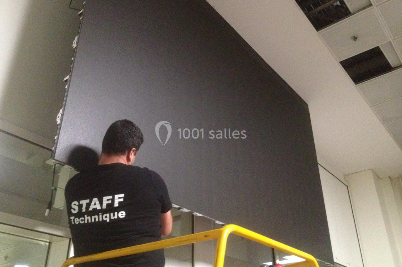 Un technicien installe un grand écran noir sur un mur, debout sur une plateforme élévatrice dans un espace intérieur.
