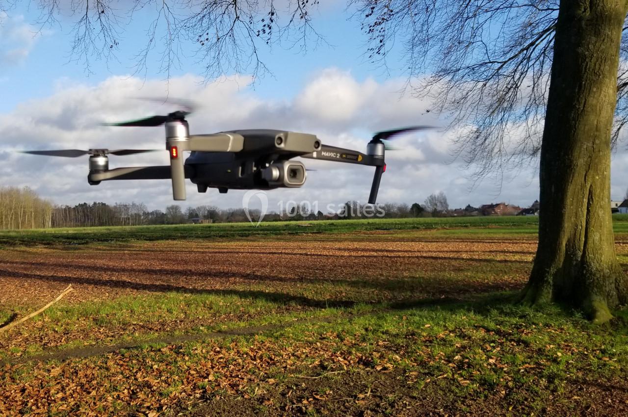 Un drone gris en vol stationnaire au-dessus d'un champ, près d'un arbre, sous un ciel partiellement nuageux.