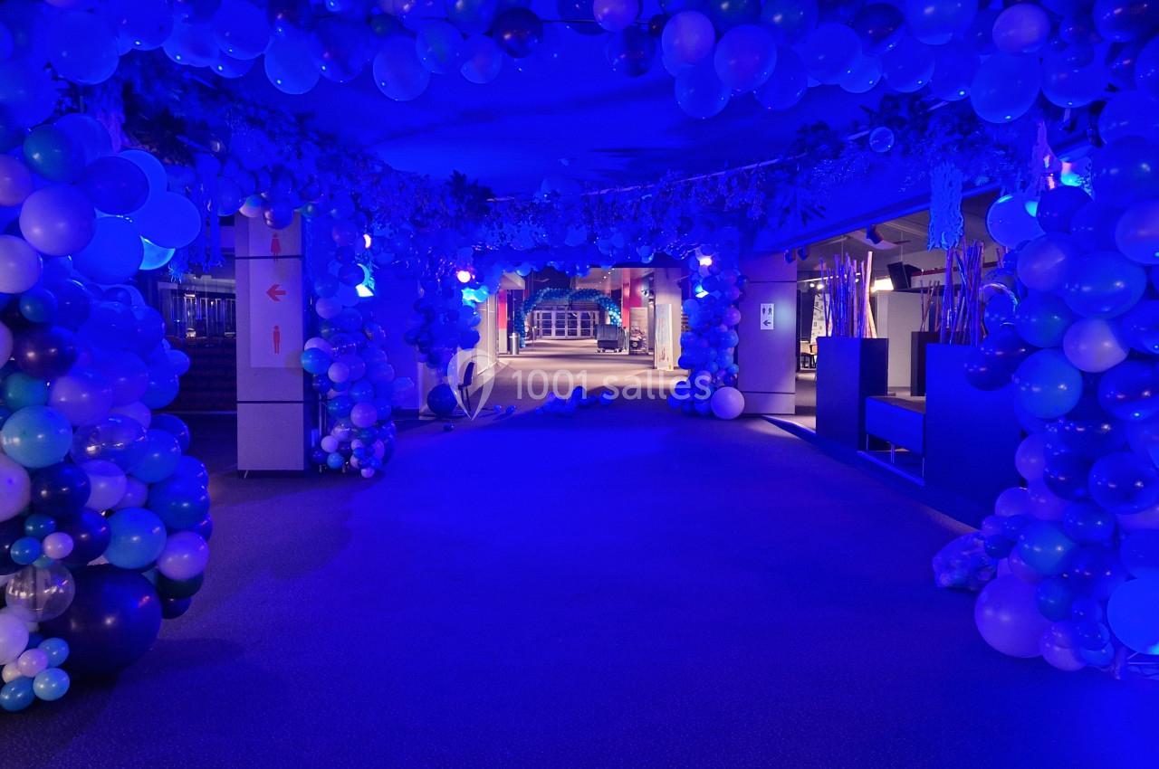 Couloir décoré de ballons bleus et blancs éclairé par des lumières bleues, créant une ambiance festive.