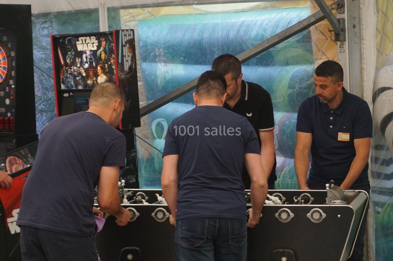 Quatre hommes jouent au baby-foot dans un espace couvert, avec une borne d'arcade visible en arrière-plan.