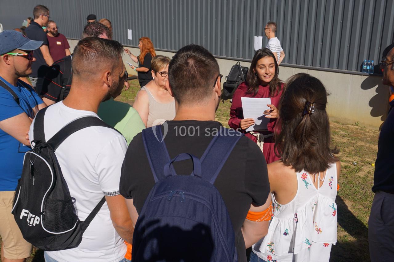Une femme s'adresse à un groupe de personnes en extérieur, près d'un bâtiment gris, sous un ciel ensoleillé.