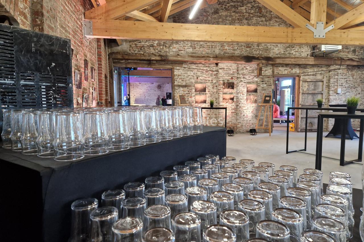 Salle en briques avec poutres apparentes, tables hautes et rangées de verres empilés sur une table noire.