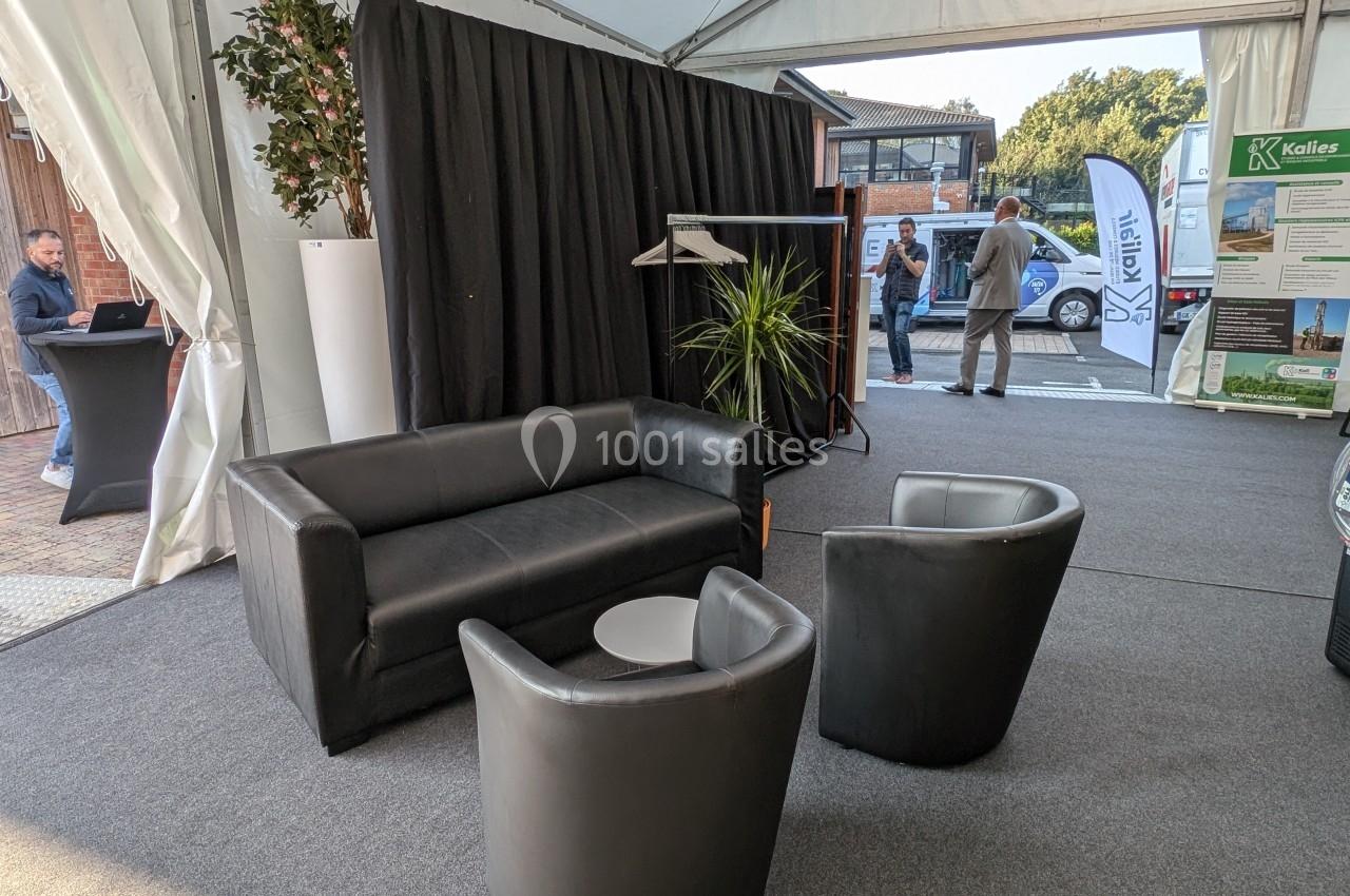 Espace lounge sous une tente avec canapé, fauteuils noirs, table basse, plantes et portants à vêtements.