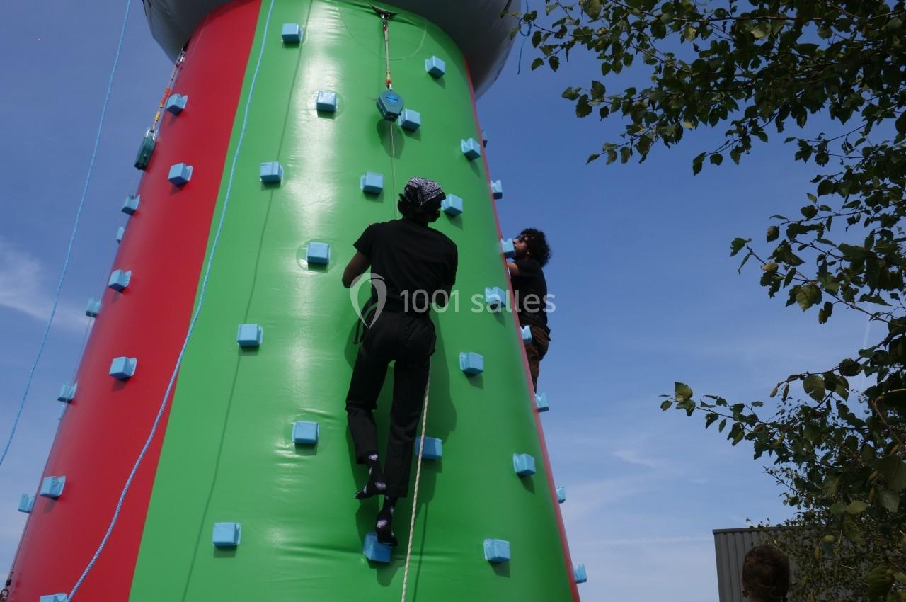 Deux personnes escaladent une structure gonflable colorée sous un ciel bleu, entourée de quelques arbres.