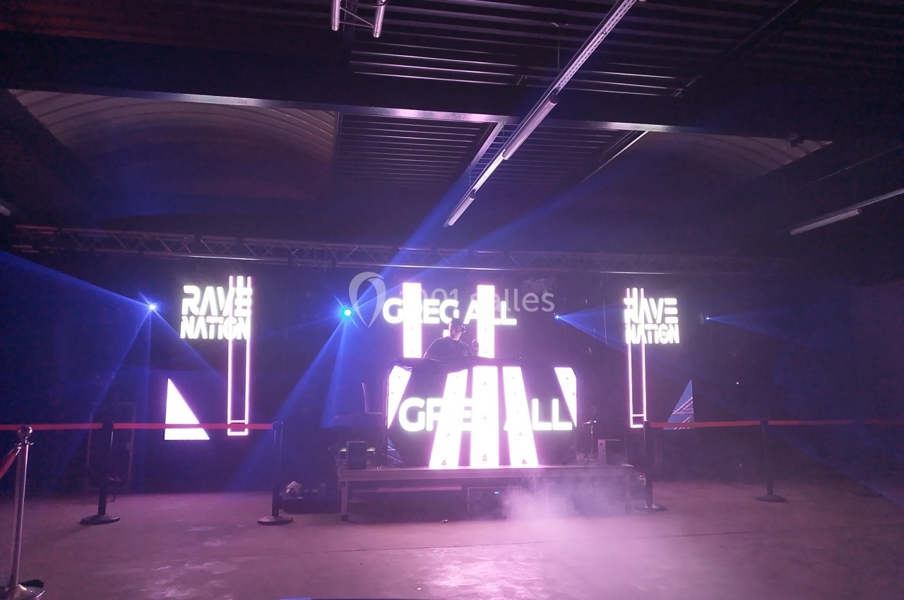 Scène de concert avec éclairages lumineux et écrans affichant des visuels, dans une salle sombre et industrielle.