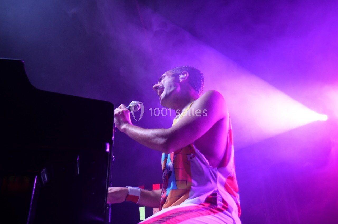 Un chanteur joue du piano sur scène sous des lumières violettes et un projecteur lumineux.