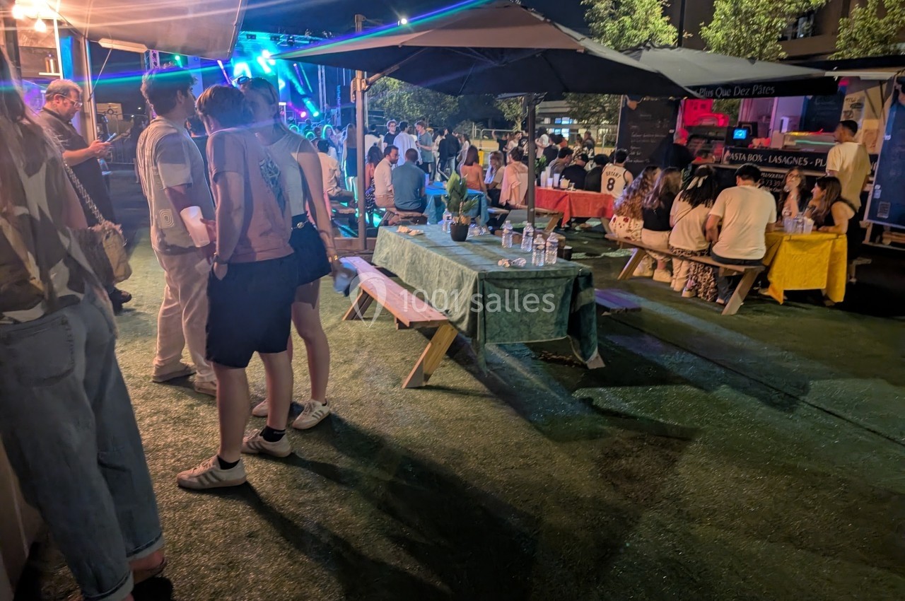 Des personnes assises et debout dans un espace extérieur éclairé, avec tables, parasols et stands en arrière-plan.