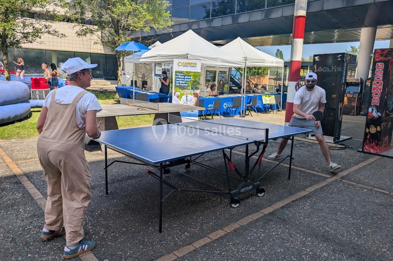 Deux personnes jouent au ping-pong en extérieur près de stands et d'une structure gonflable.