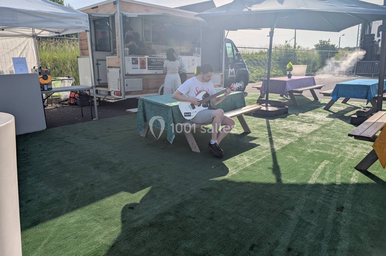 Un homme joue de la guitare assis sur un banc en bois dans un espace extérieur avec food trucks et parasols.