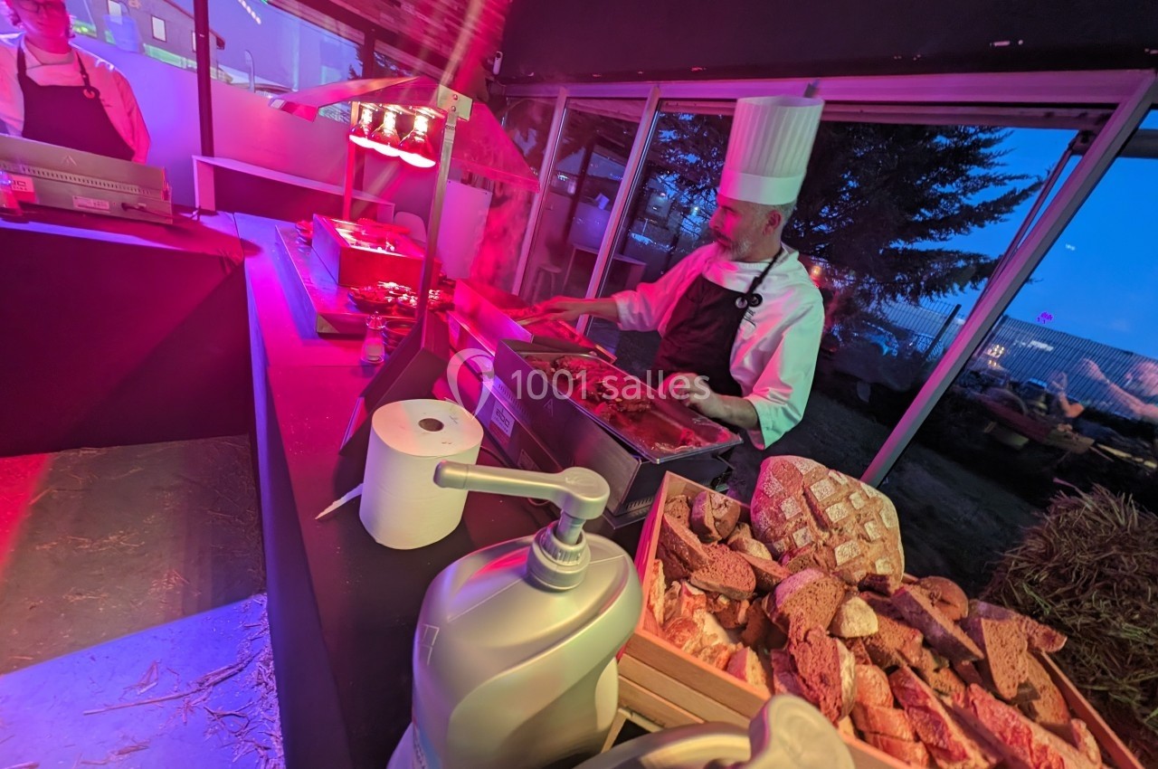 Un chef en tenue blanche prépare des plats chauds derrière un buffet avec du pain et des condiments.
