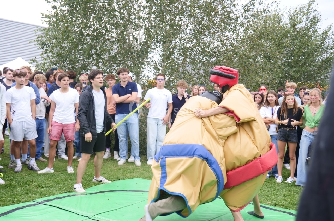 Deux personnes en costumes de sumo s'affrontent sur un tapis vert, entourées par un groupe de spectateurs en plein air.