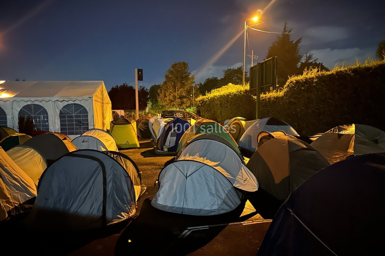 Alignement de tentes installées sur un parking éclairé la nuit, avec une grande tente blanche en arrière-plan.