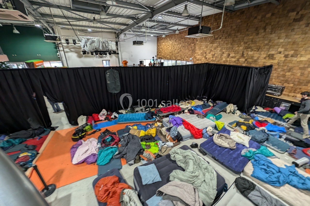 Des matelas et sacs de couchage dispersés dans une grande salle intérieure avec des rideaux noirs en fond.