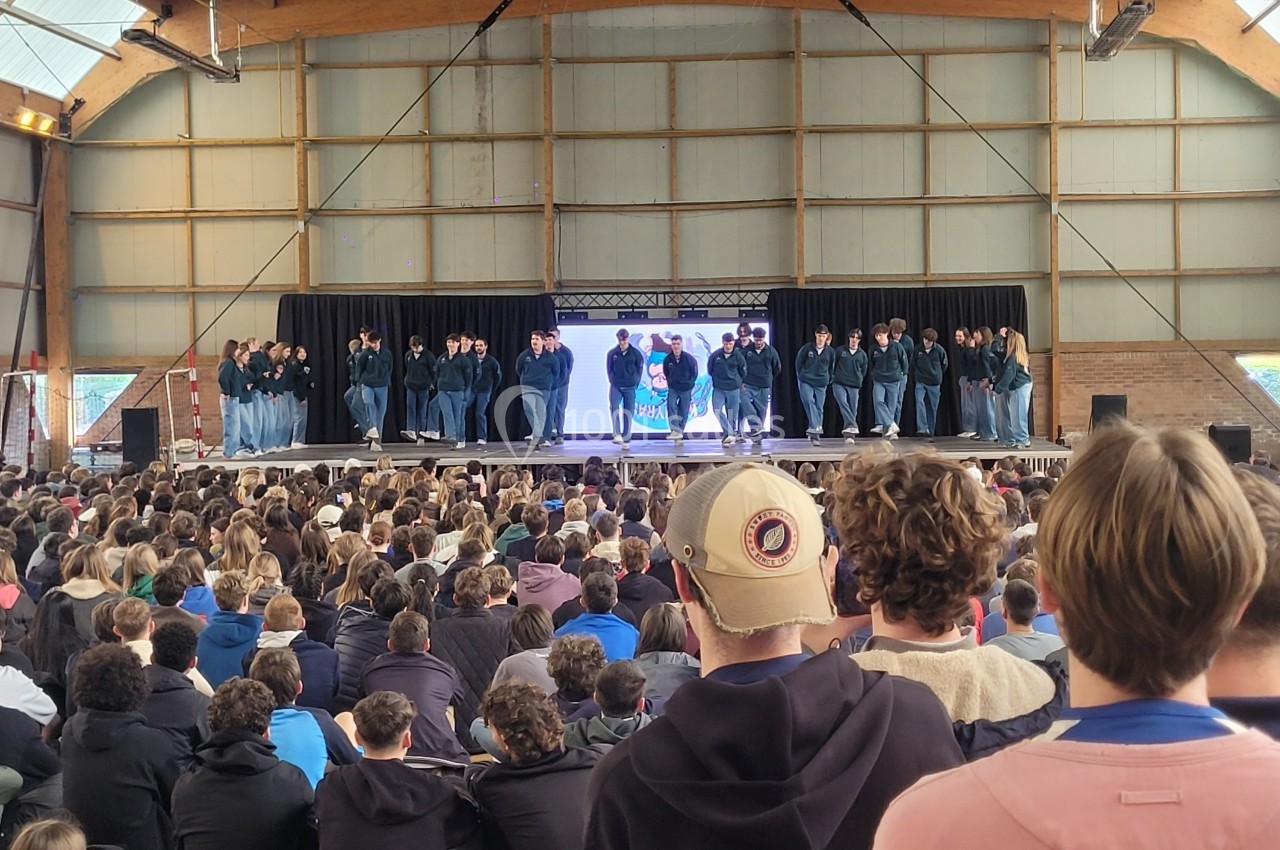 Un groupe de danseurs sur scène devant un large public dans une salle avec une structure en bois.