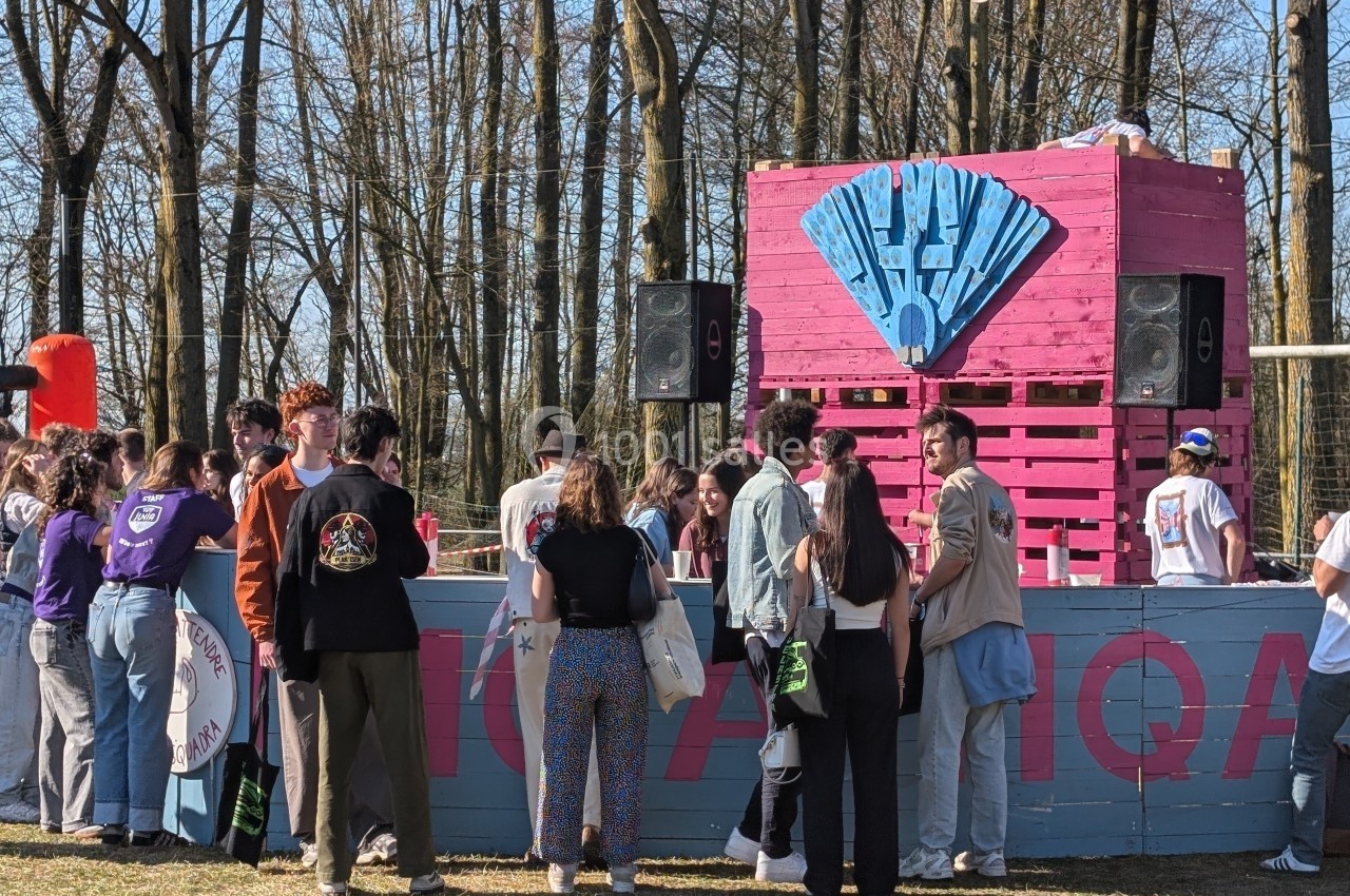 Des personnes rassemblées devant un stand en bois rose avec des enceintes, dans un espace extérieur boisé.