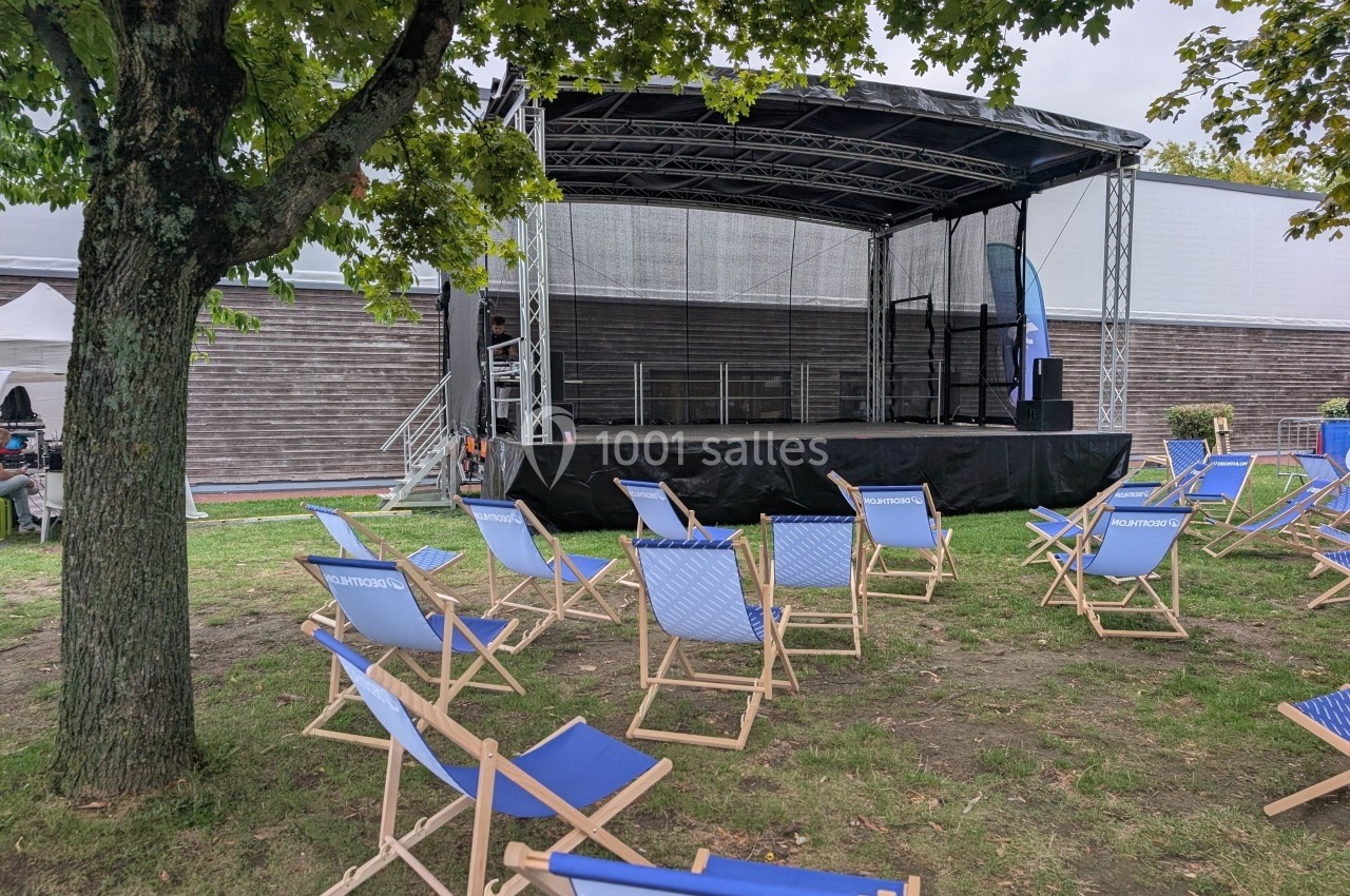 Scène extérieure vide avec des chaises pliantes bleues disposées sur une pelouse sous un arbre.