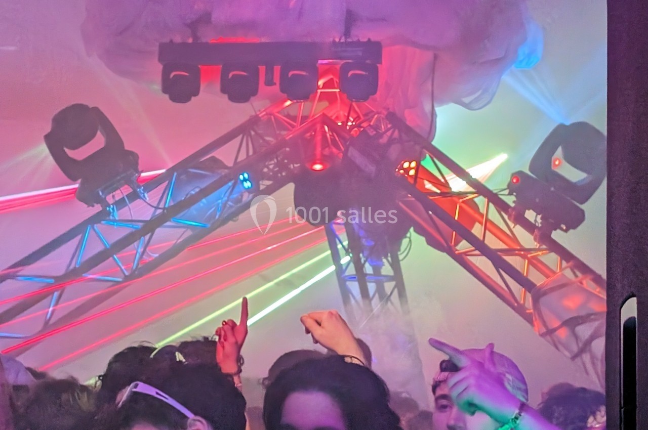 Foule dans une salle de concert avec des jeux de lumière colorés et une structure métallique au plafond.