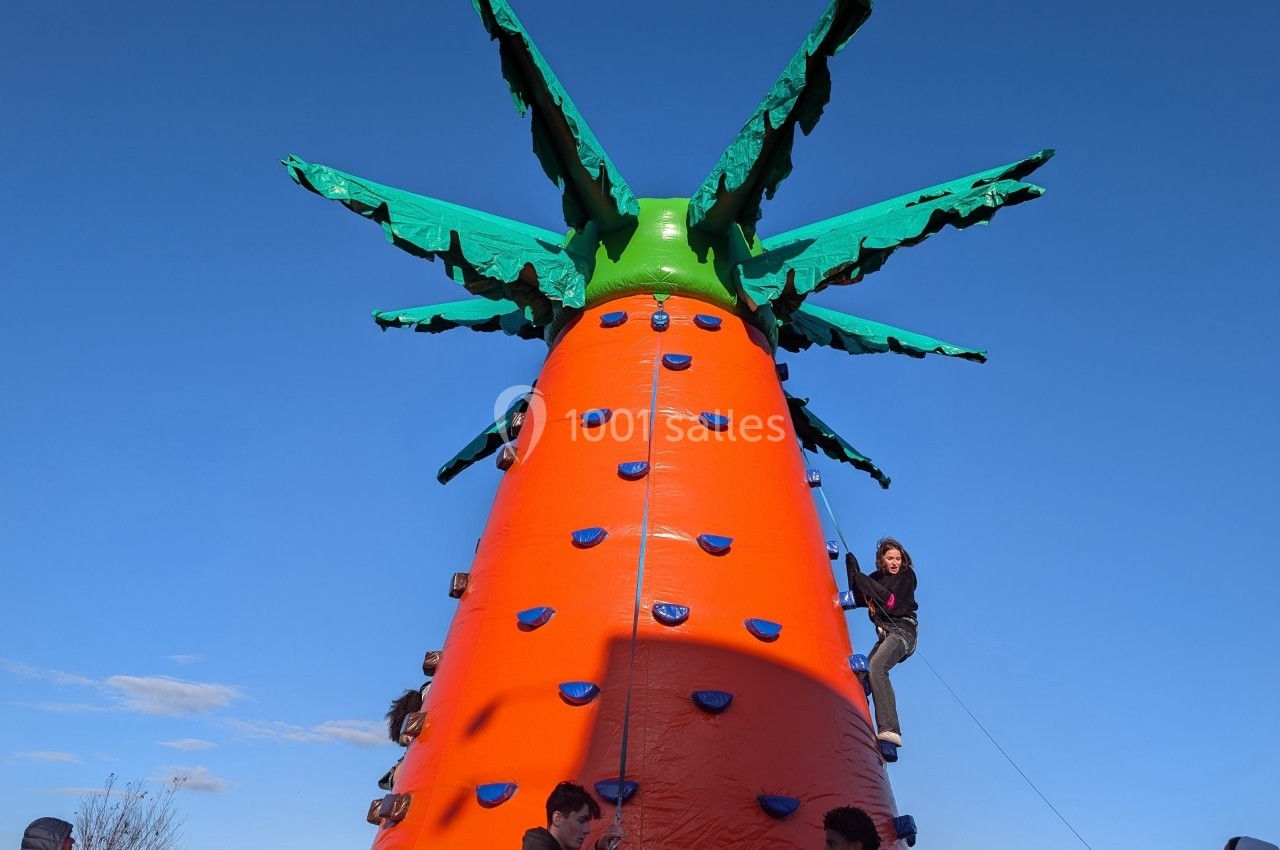 Structure gonflable en forme de carotte géante avec des enfants escaladant ses prises colorées sous un ciel bleu.