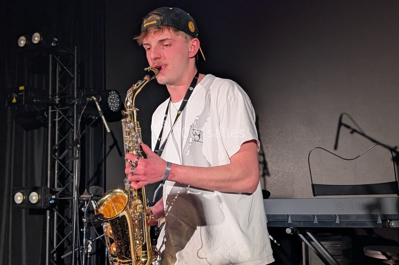 Un musicien joue du saxophone sur une scène sombre, entouré d'équipements de concert.