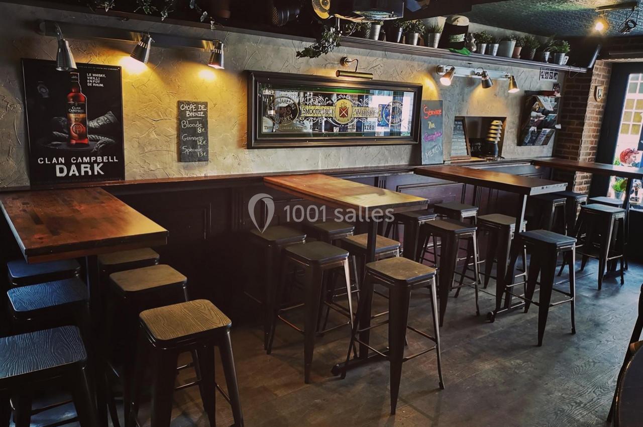 Intérieur d'un bar avec tables hautes en bois, tabourets métalliques et décorations murales dans une ambiance tamisée.