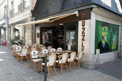 Terrasse de café avec des chaises en osier, tables vides et parasols, située dans une rue pavée bordée de bâtiments.