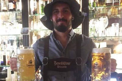 Terrasse de café avec des chaises en osier, tables vides et parasols, située dans une rue pavée bordée de bâtiments.