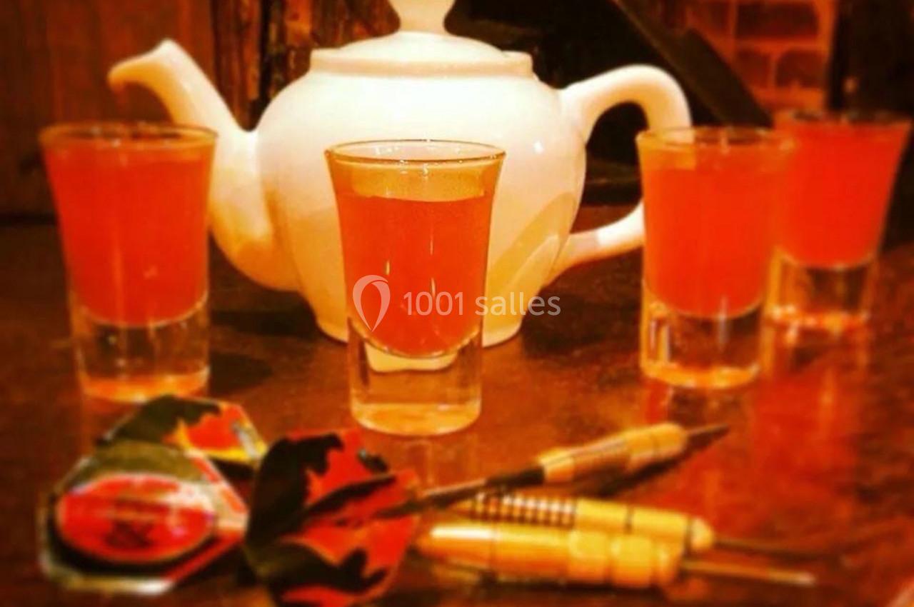 Théière blanche entourée de quatre verres de liquide orange, avec des fléchettes posées au premier plan.