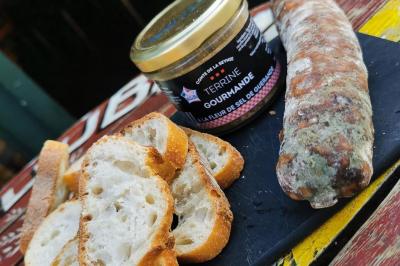 Terrasse de café avec des chaises en osier, tables vides et parasols, située dans une rue pavée bordée de bâtiments.