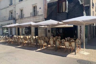 Terrasse de café avec des chaises en osier, tables vides et parasols, située dans une rue pavée bordée de bâtiments.