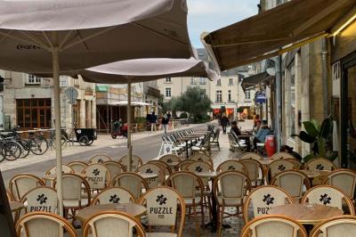 Terrasse de café avec des chaises en osier, tables vides et parasols, située dans une rue pavée bordée de bâtiments.