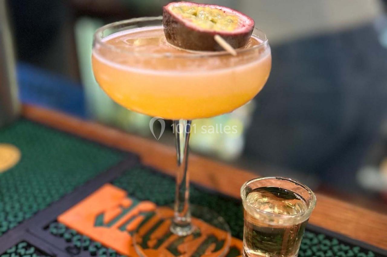 Cocktail dans une coupe décorée d'un demi-fruit de la passion, accompagné d'un petit verre de liqueur sur un bar.