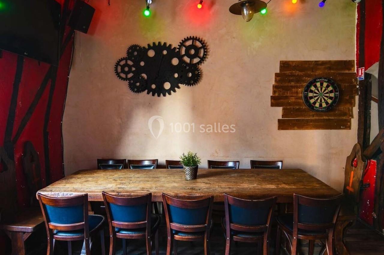 Salle avec une grande table en bois entourée de chaises, décorée de guirlandes lumineuses et d'engrenages au mur.