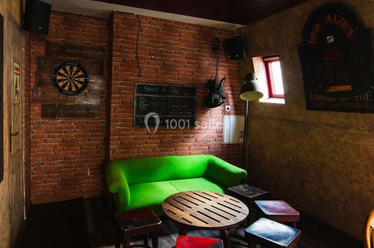 Salle avec murs en briques, canapé vert, table basse en bois, tabourets, cible de fléchettes et tableau noir.