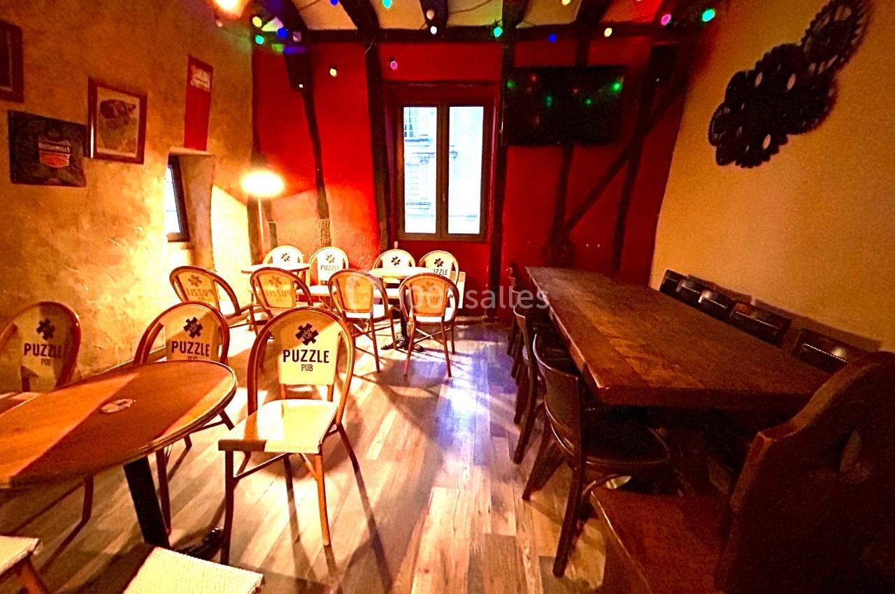 Salle chaleureuse avec murs en pierre, tables en bois, chaises variées et guirlandes lumineuses colorées.