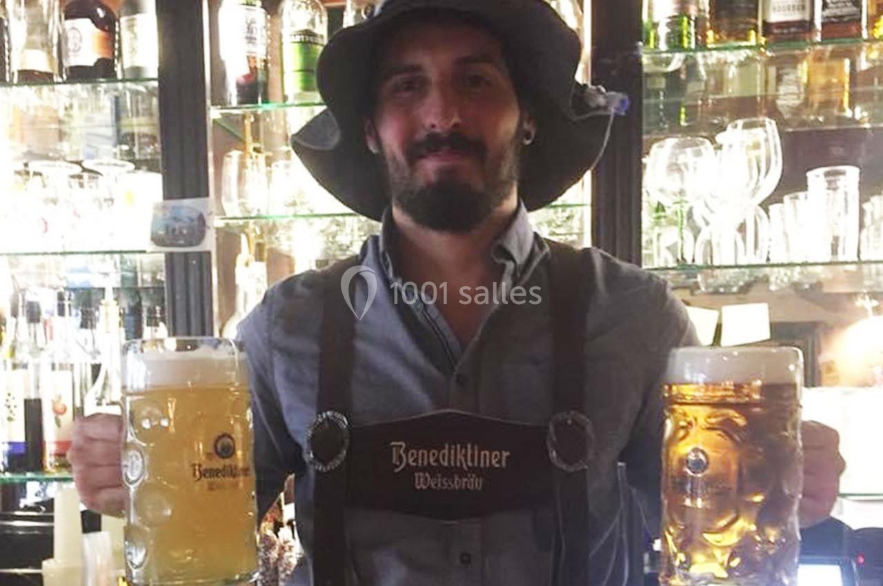 Un homme en tenue traditionnelle tient deux chopes de bière dans un bar avec des bouteilles en arrière-plan.
