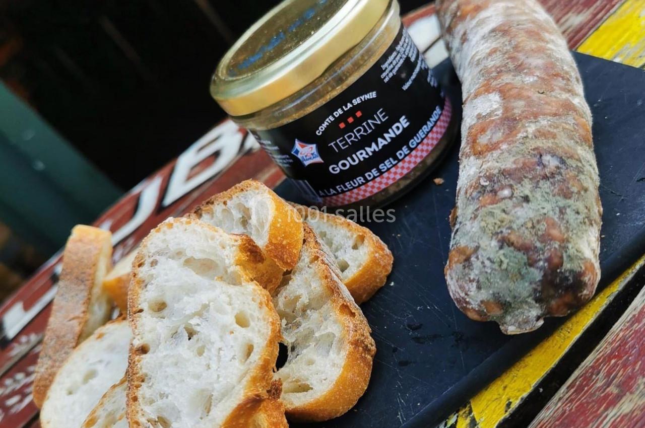 Tranches de pain, terrine en pot et saucisson disposés sur une planche noire en bois.