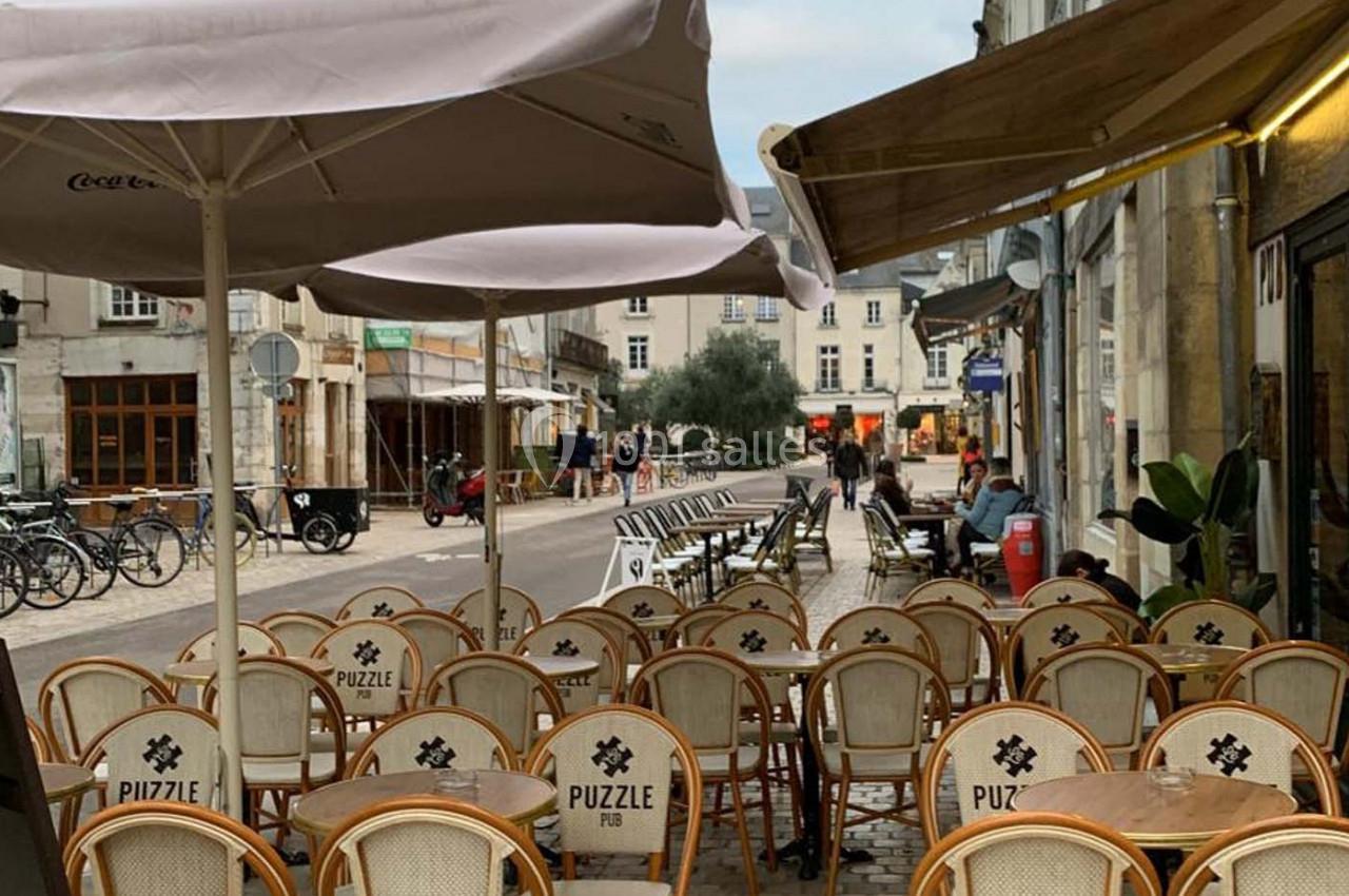 Terrasse d'un café avec des chaises en osier, tables vides et parasols, située dans une rue calme bordée de bâtiments.
