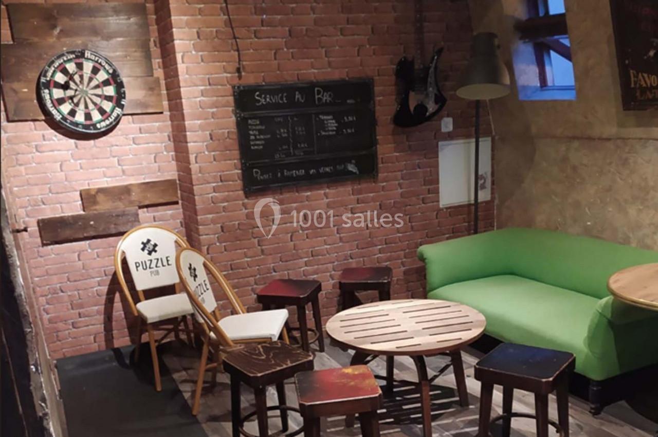 Espace intérieur avec murs en briques, cible de fléchettes, chaises, tables en bois et canapé vert.