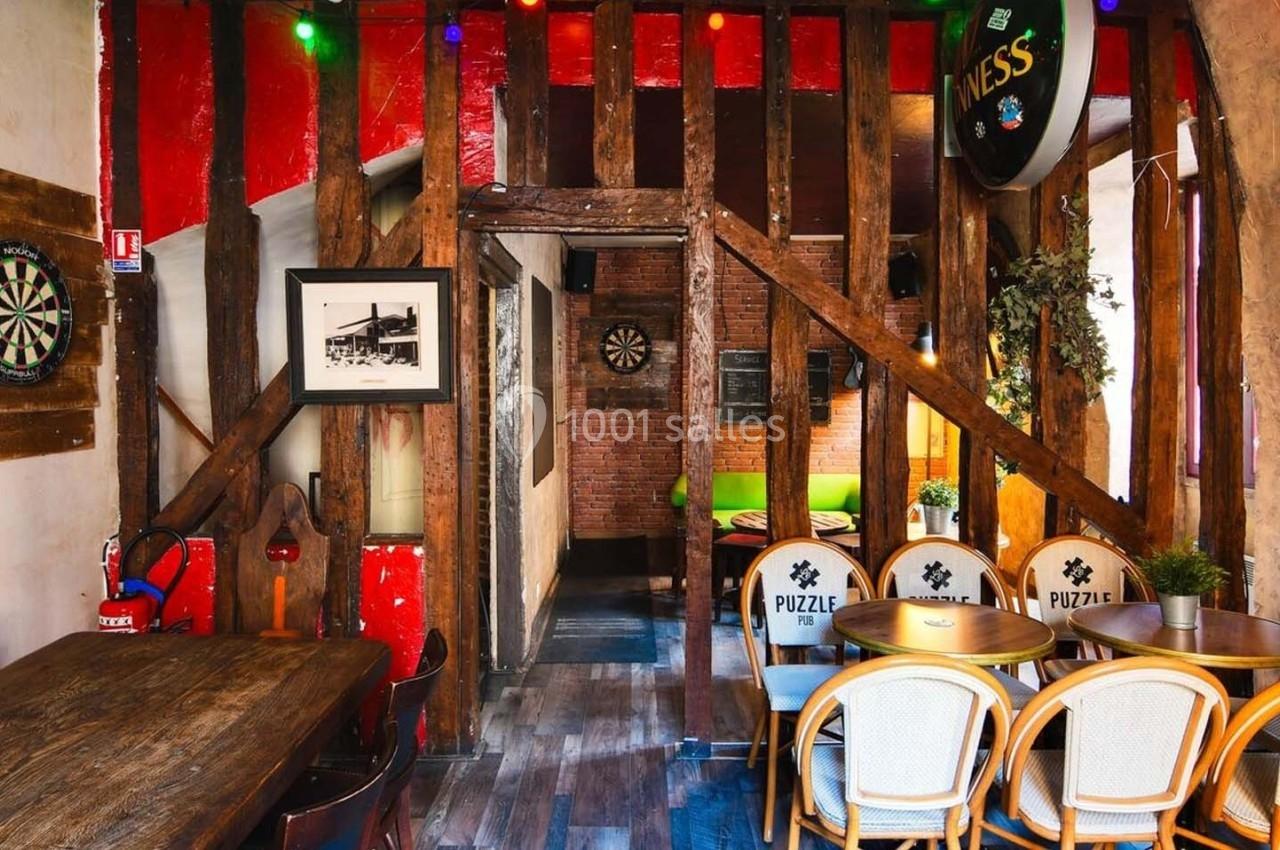 Salle conviviale avec murs en bois et briques, tables en bois, chaises en rotin, cible de fléchettes et décoration lumineuse.