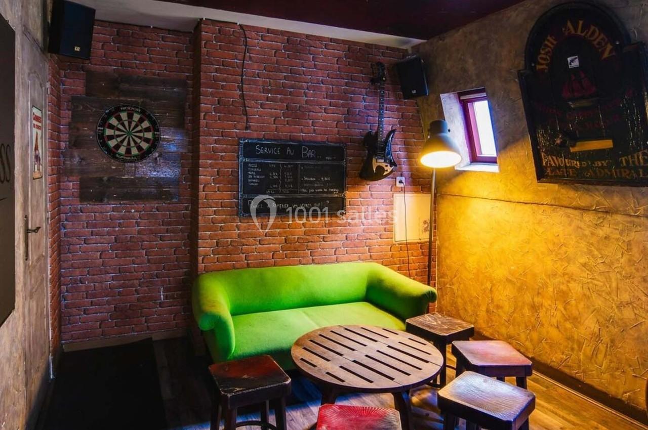 Salle conviviale avec murs en briques, canapé vert, table basse, tabourets, cible de fléchettes et éclairage tamisé.