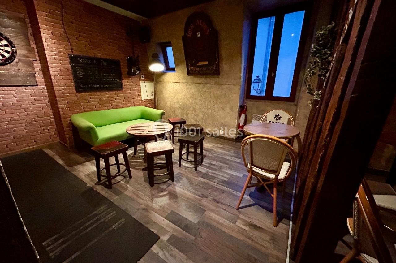 Salle avec murs en briques, canapé vert, chaises et tables en bois, ambiance tamisée et fenêtre donnant sur l'extérieur.