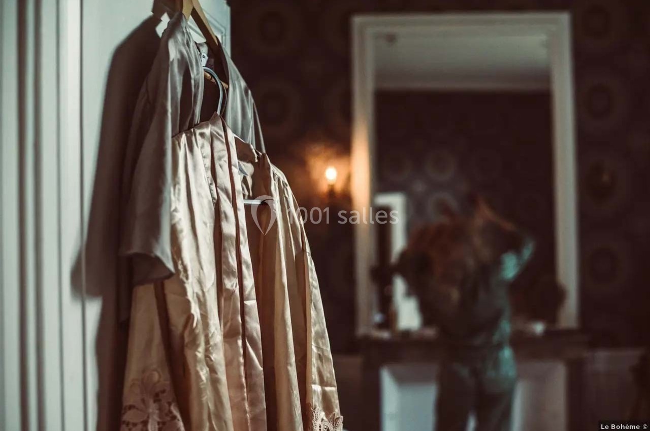 Robe beige suspendue à un cintre au premier plan, avec une personne floue se coiffant devant un miroir en arrière-plan.