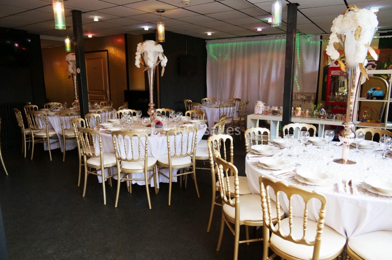Salle de réception décorée avec des tables rondes dressées, chaises dorées et centres de table ornés de plumes blanches.