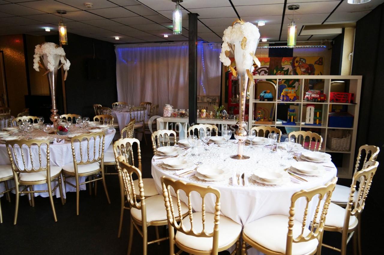 Salle de réception décorée avec des tables rondes dressées, chaises dorées et centres de table ornés de fleurs blanches.