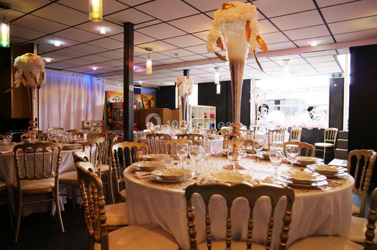 Salle de réception décorée avec des tables rondes, nappes blanches, chaises dorées et centres de table floraux.